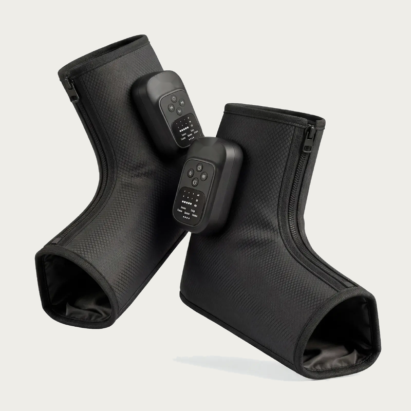 NACRE FORM™ cordless air compression foot and ankle massager displayed as a pair of black wearable massage boots against a warm cream background. Each electric foot massager features a textured fabric exterior, integrated digital control panel, adjustable massage functions and front zip fastening. The product photograph presents a rechargeable heated foot massage device with comfortable ankle coverage, portable construction and personalised compression settings for convenient home relaxation.
