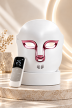 Premium white LED light therapy face mask with metallic rose-gold detailing, displayed on its storage stand alongside a wireless remote control with a digital 25-minute timer. This rechargeable facial light therapy mask features adjustable LED colour settings and a modern design for an at-home skincare and beauty routine.
