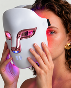 Woman holding a white LED red light therapy face mask with elegant muted rose-gold metallic detailing during her at-home skincare routine. The rechargeable facial light therapy mask features illuminated eye openings, a comfortable wearable design and multiple LED light colours for a modern beauty and self-care experience.
