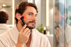 Man wearing a cream bathrobe using the NACRE FORM™ handheld red light therapy wand on his cheek and jawline during an at-home facial skincare routine in a modern bathroom. The lifestyle image shows a black LED facial beauty device with a glowing red light treatment head positioned against the side of his face, demonstrating targeted application around the cheek and jawline. This compact red light face wand is presented as a skincare tool for men interested in red light therapy for the face, facial grooming