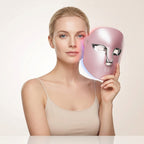 Blonde woman holding a pink LED red light therapy face mask beside her face against a soft neutral background. The NACRE FORM™ rechargeable LED facial beauty device features a metallic silver eye and nose design, an ergonomic full-face shape and four light settings, including red, blue, pink and near-infrared. Designed for convenient at-home skincare, this LED light therapy face mask complements beauty routines focused on the appearance of fine lines, uneven skin texture and visible signs of ageing.